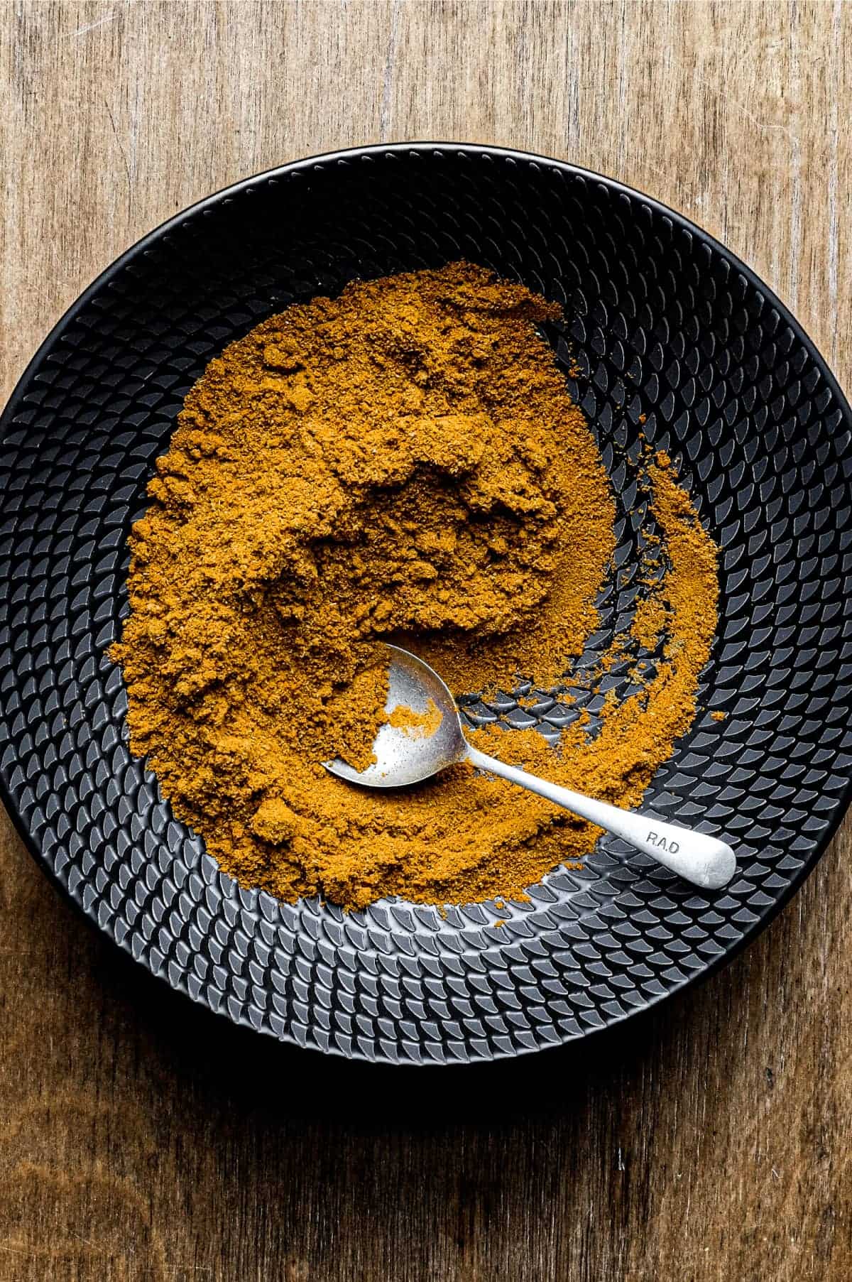 A black bowl of Madras Curry Powder on a wooden surface with a small spoon.