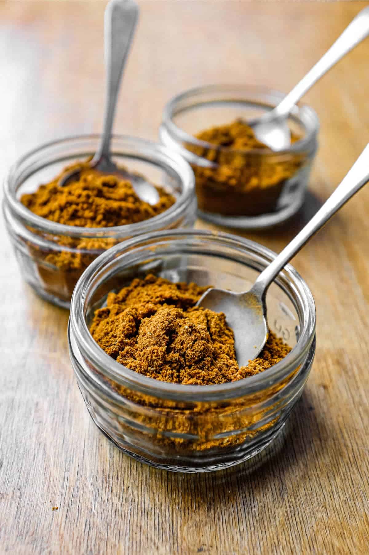 Three small jars of Madras curry powder with spoons on a wooden surface