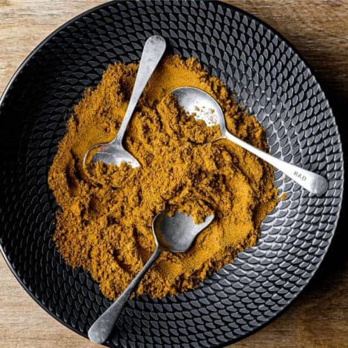 A black bowl of Madras Curry Powder on a wooden surface with three small spoons.