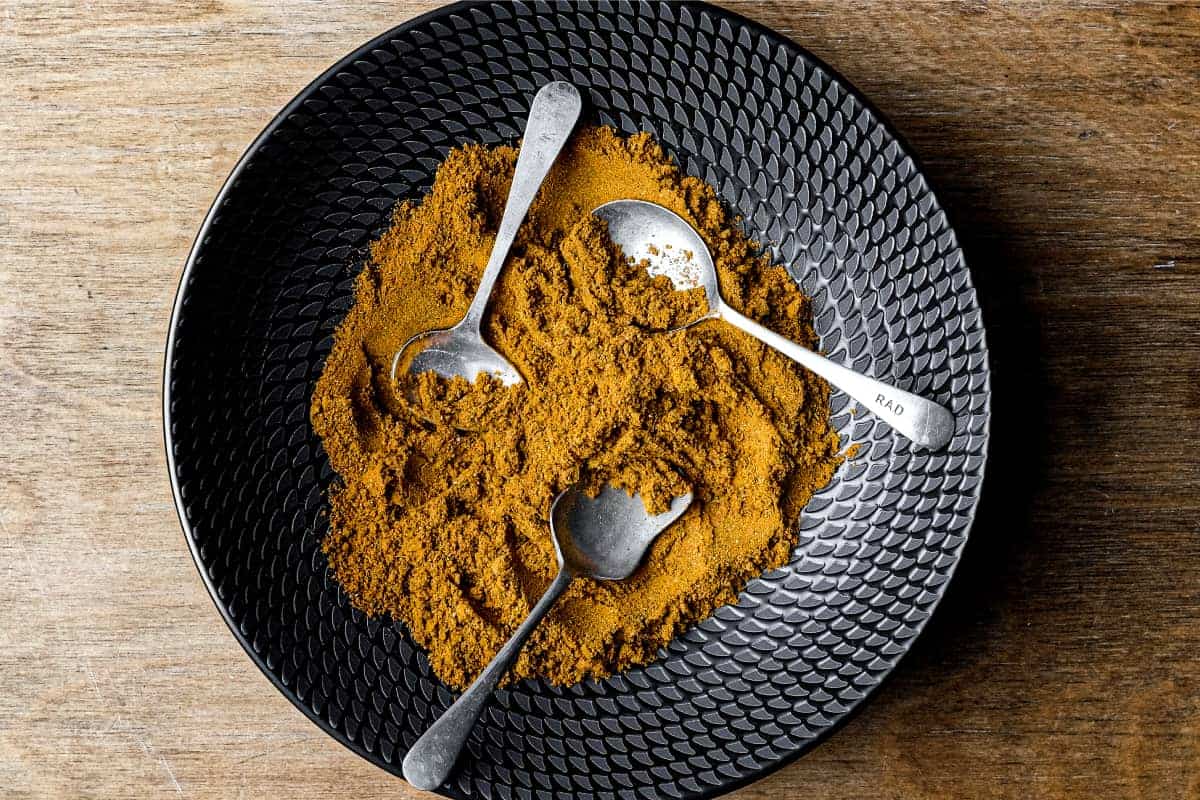 A black bowl of Madras Curry Powder on a wooden surface with three small spoons.