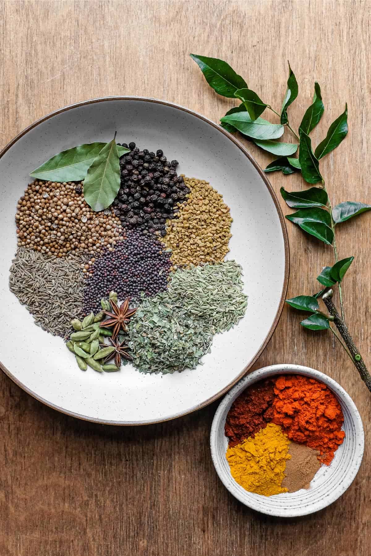 The ingredients of whole and ground spices plus a branch of fresh curry leaves on a wooden surface.