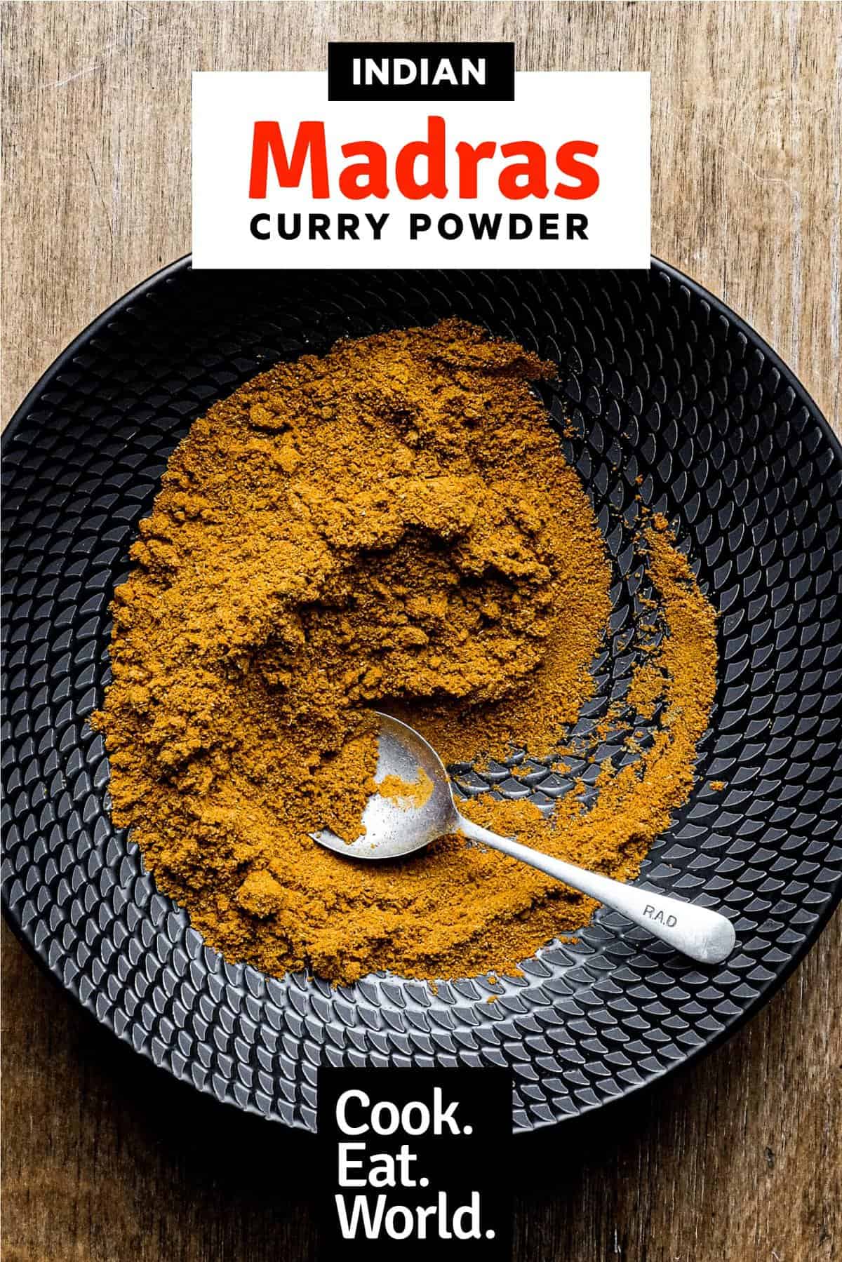 A black bowl of Madras Curry Powder on a wooden surface with a small spoon.