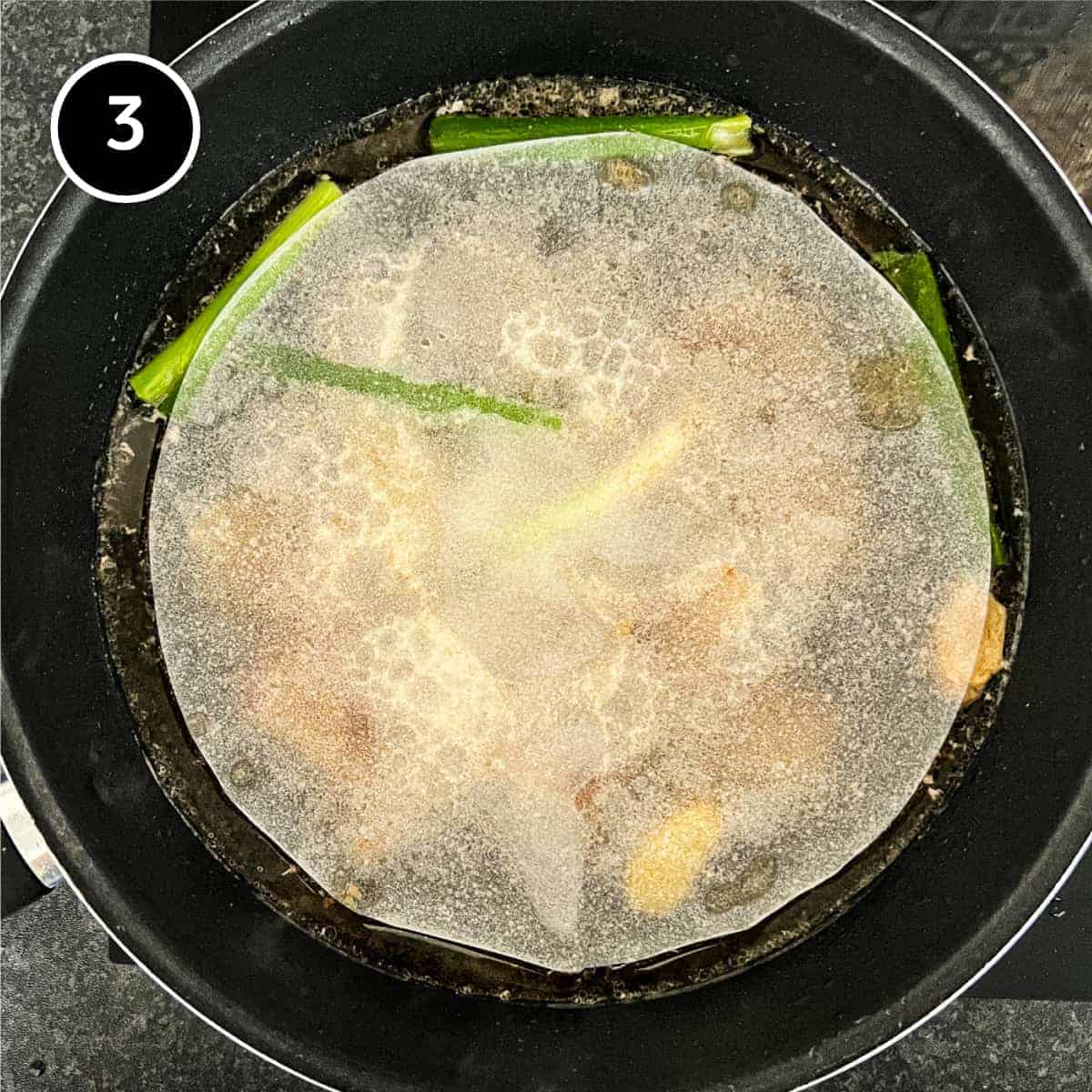 a sheet of parchment paper covers the contents of a pan of simmering pork belly