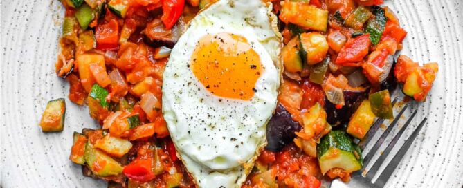 A plate of Spanish Pisto Manchego topped with a crisp fried egg. A fork sits alongside.