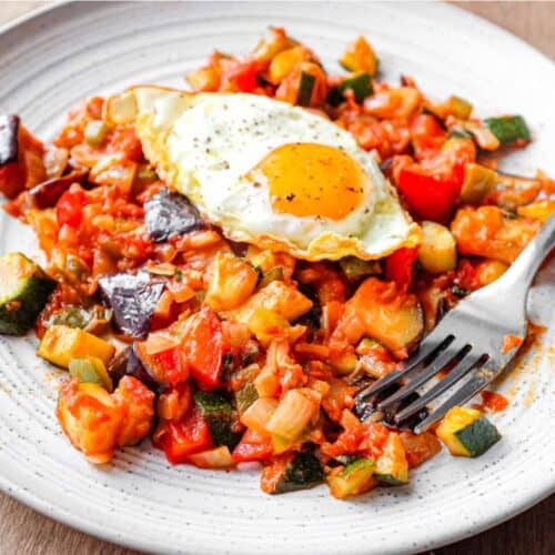 A plate of Spanish Pisto Manchego topped with a crisp fried egg. A fork sits alongside.