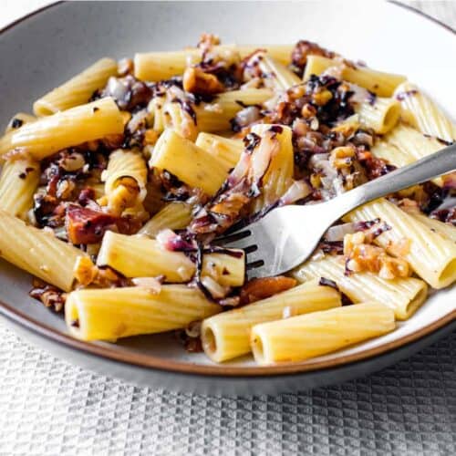 A bowl of rigatoni pasta with a radicchio, walnut and guanciale sauce in a bowl with a fork