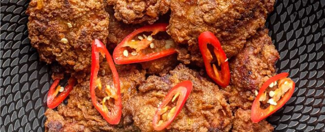 A black bowl filled with Malaysian fried chicken, Ayam Goreng, garnished with red chillies and accompanied by lime wedges.