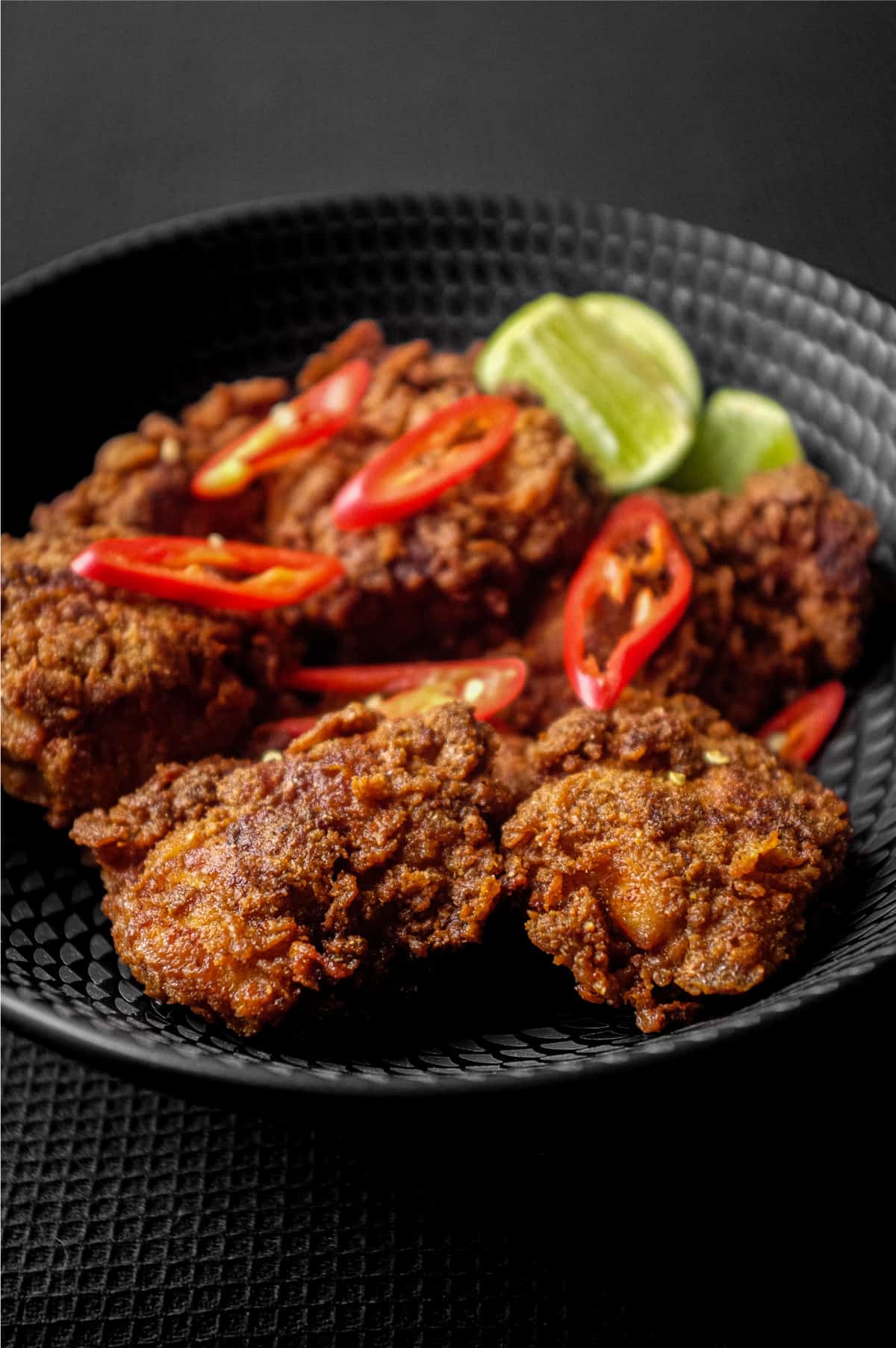 A black bowl filled with Malaysian fried chicken, Ayam Goreng, garnished with red chillies and accompanied by lime wedges.