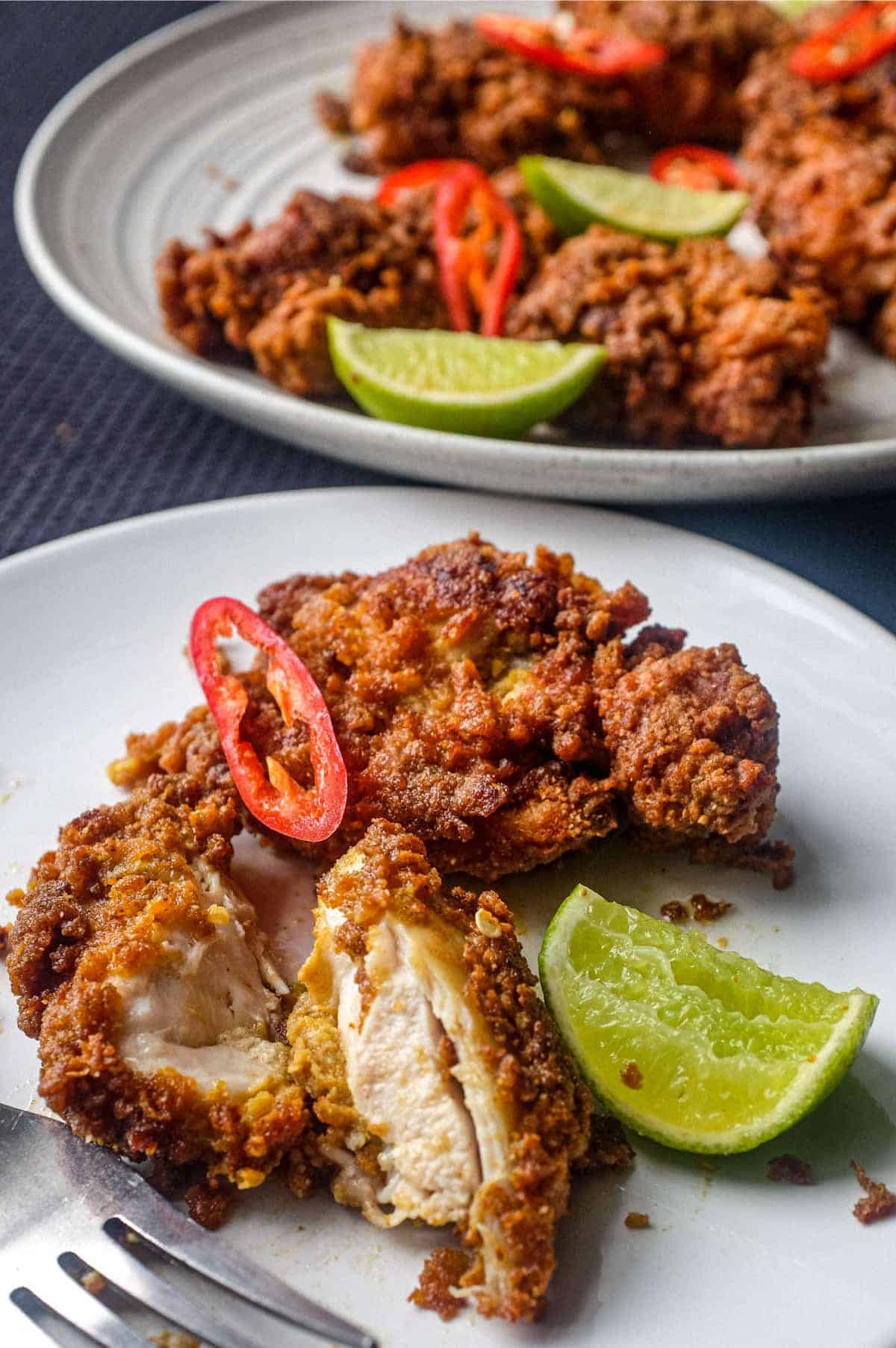 A piece of Ayam Goreng (Malaysian fried chicken) on plate with a lime wedge and slice of chilli. A bowl of the chicken sits in the background.