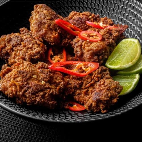 A black bowl filled with Malaysian fried chicken, Ayam Goreng, garnished with red chillies and accompanied by lime wedges.
