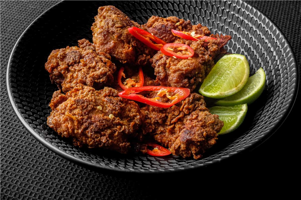 A black bowl filled with Malaysian fried chicken, Ayam Goreng, garnished with red chillies and accompanied by lime wedges.