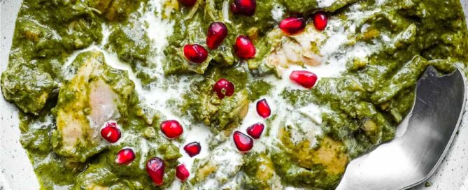 A bowl of Chicken Saag curry with a spoon, drizzled with fresh cream and sprinkled with pomegranate seeds.