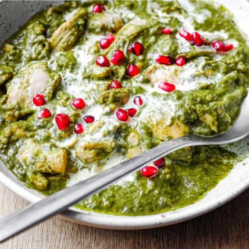 A bowl of Chicken Saag curry with a spoon, drizzled with fresh cream and sprinkled with pomegranate seeds.