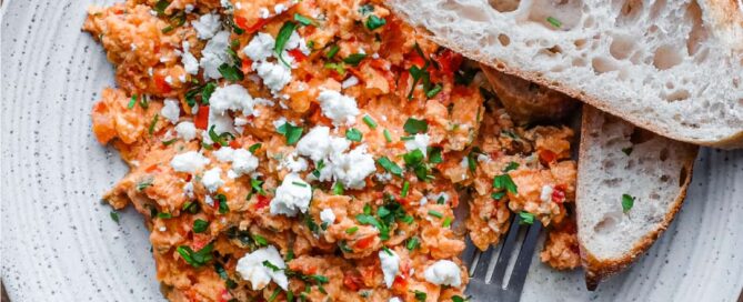 A plate of Greek Strapatsada scattered with feta cheese and parsley. Crusty bread and a fork sits alongside.