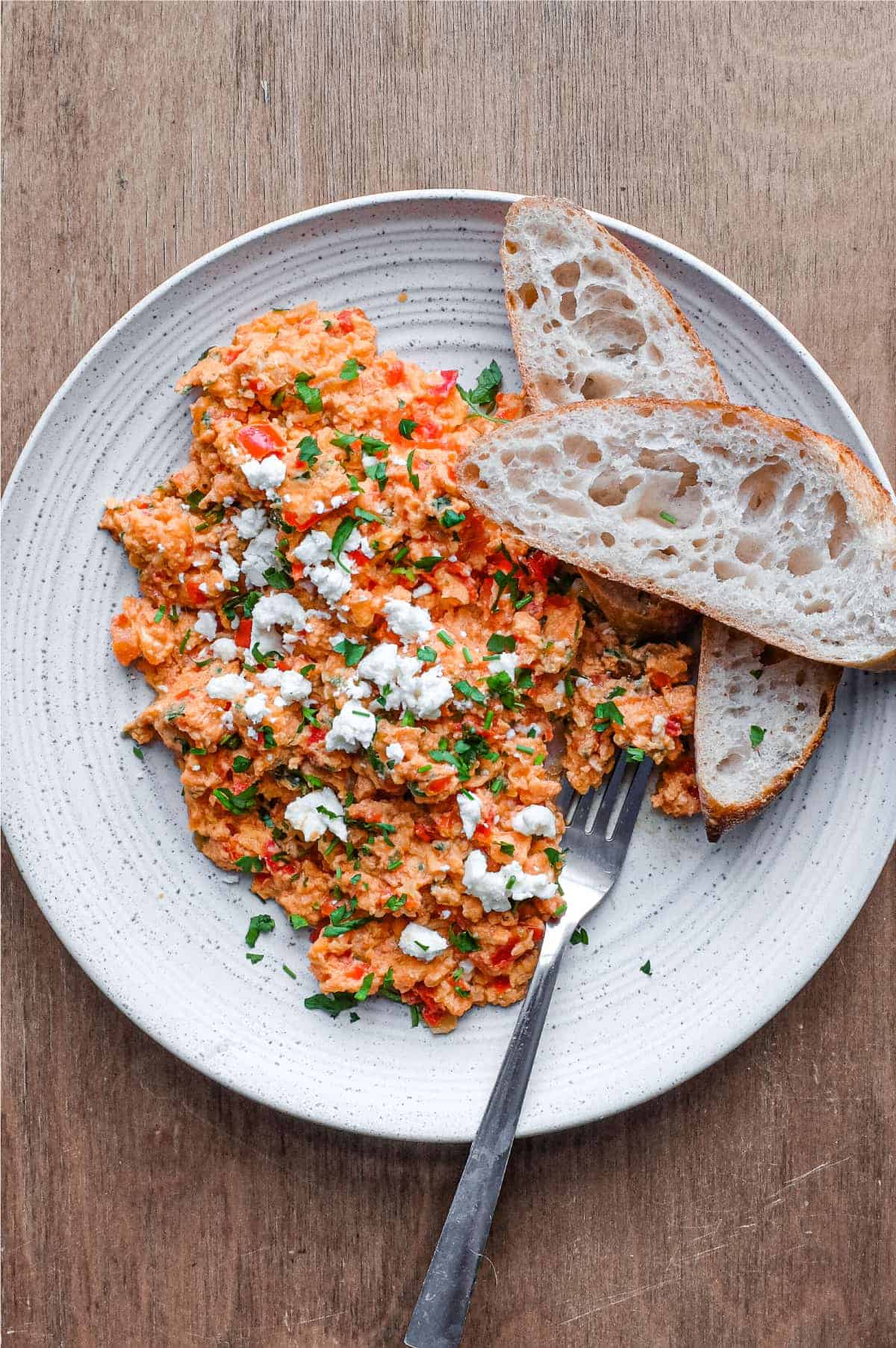 A plate of Greek Strapatsada scattered with feta cheese and parsley. Crusty bread and a fork sits alongside.