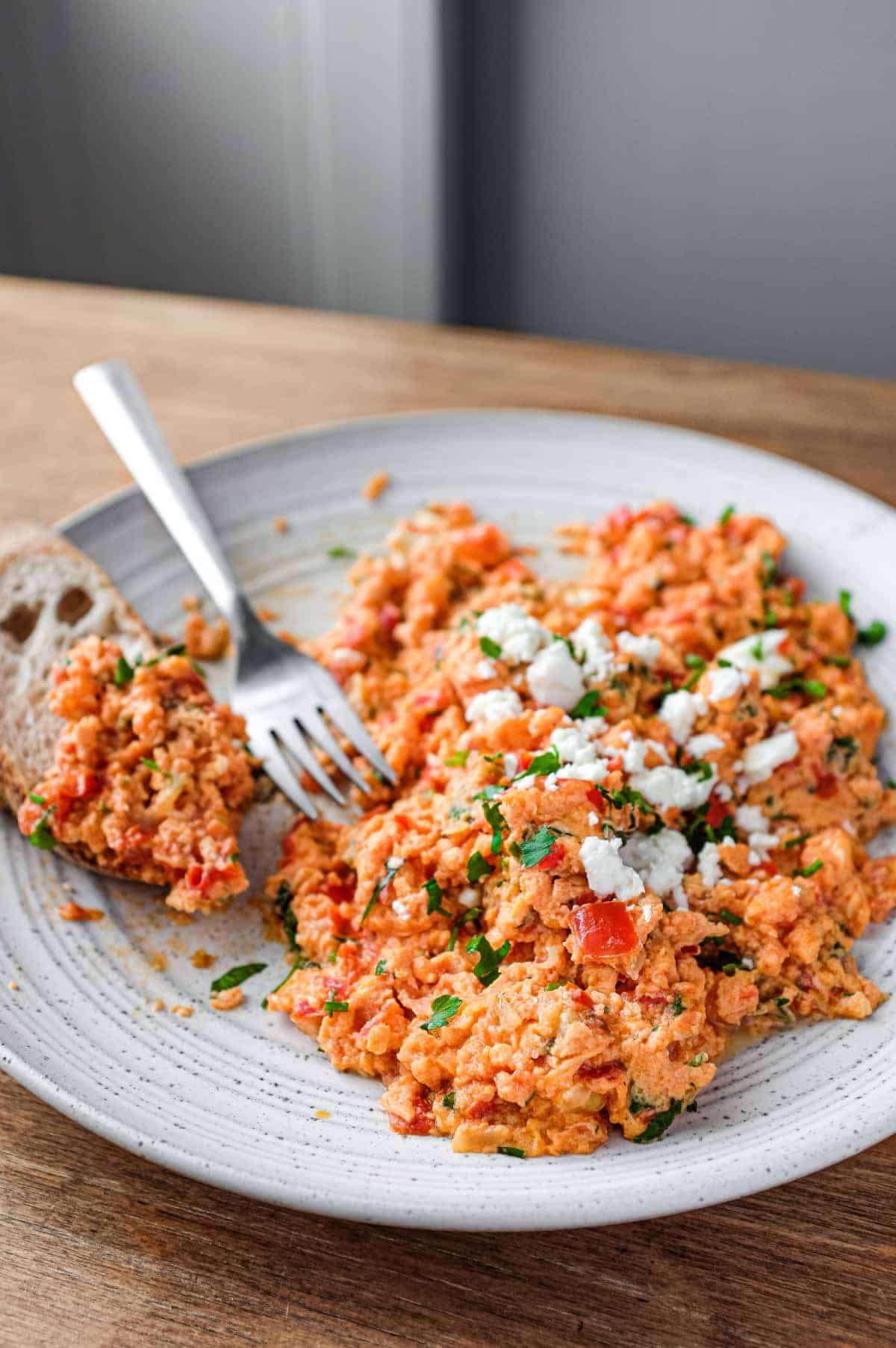 A plate of Greek Strapatsada scattered with feta cheese and parsley. Crusty bread and a fork sits alongside.