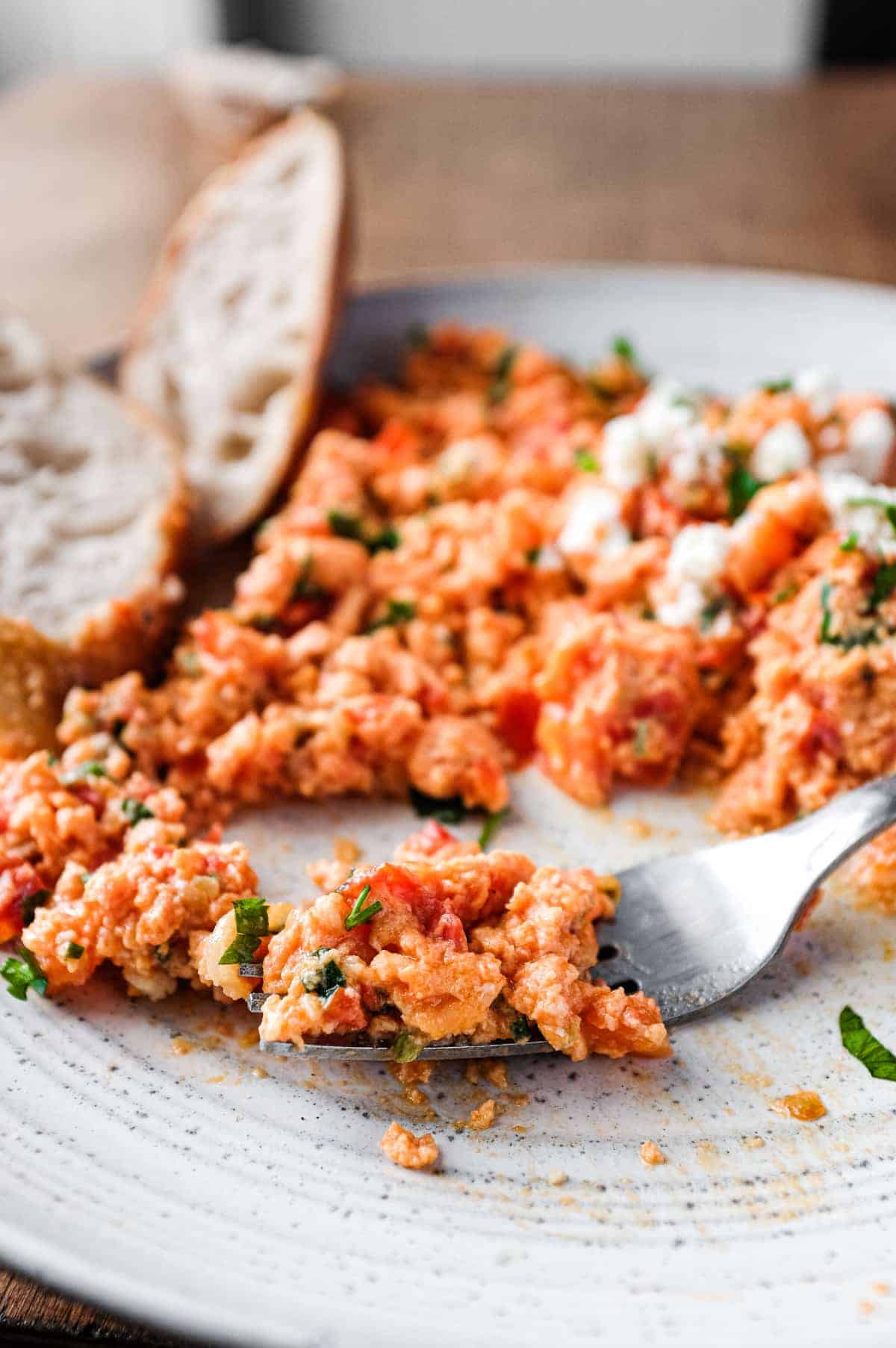 A plate of Greek Strapatsada scattered with feta cheese and parsley. Crusty bread and a fork sits alongside.