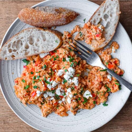 A plate of Greek Strapatsada scattered with feta cheese and parsley. Crusty bread and a fork sits alongside.