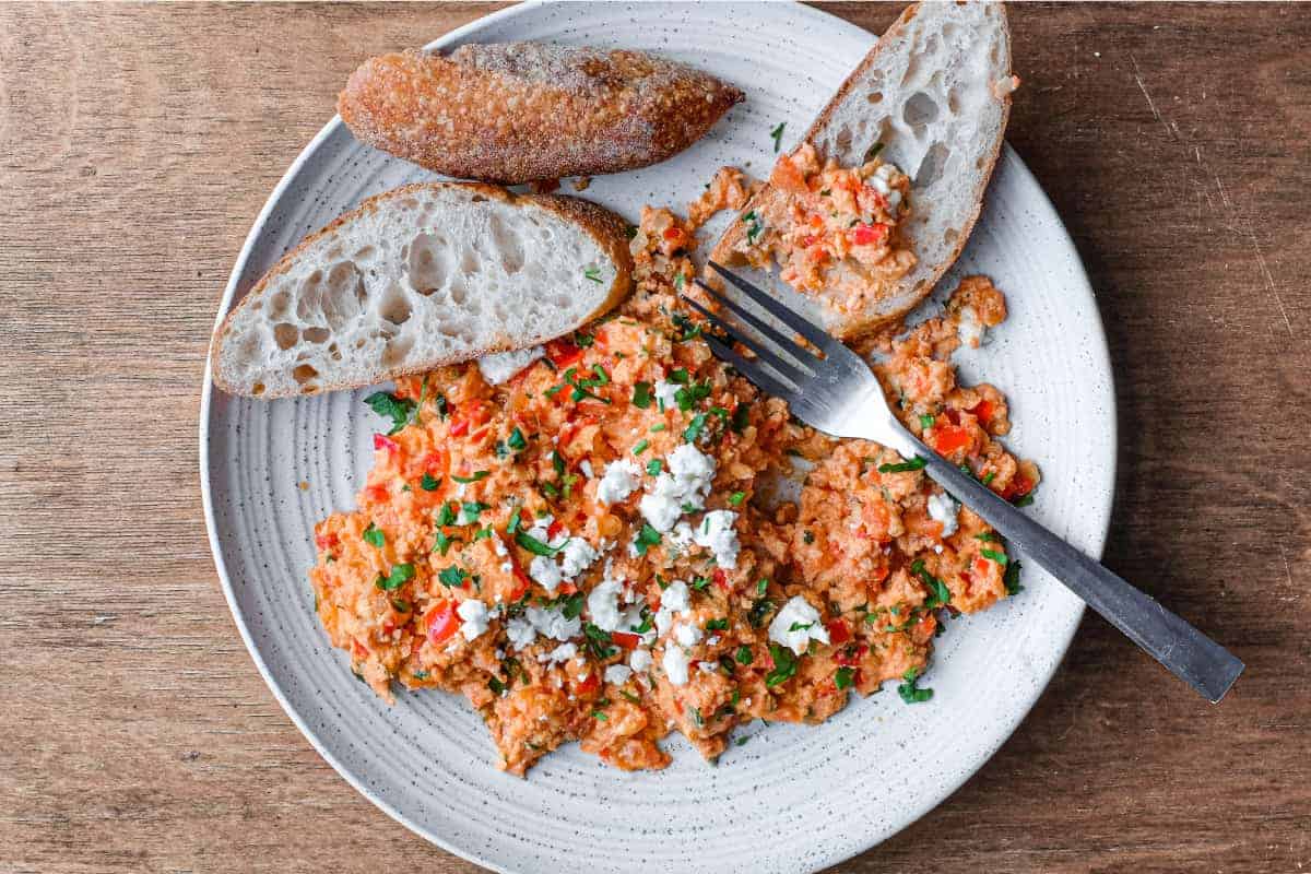 A plate of Greek Strapatsada scattered with feta cheese and parsley. Crusty bread and a fork sits alongside.