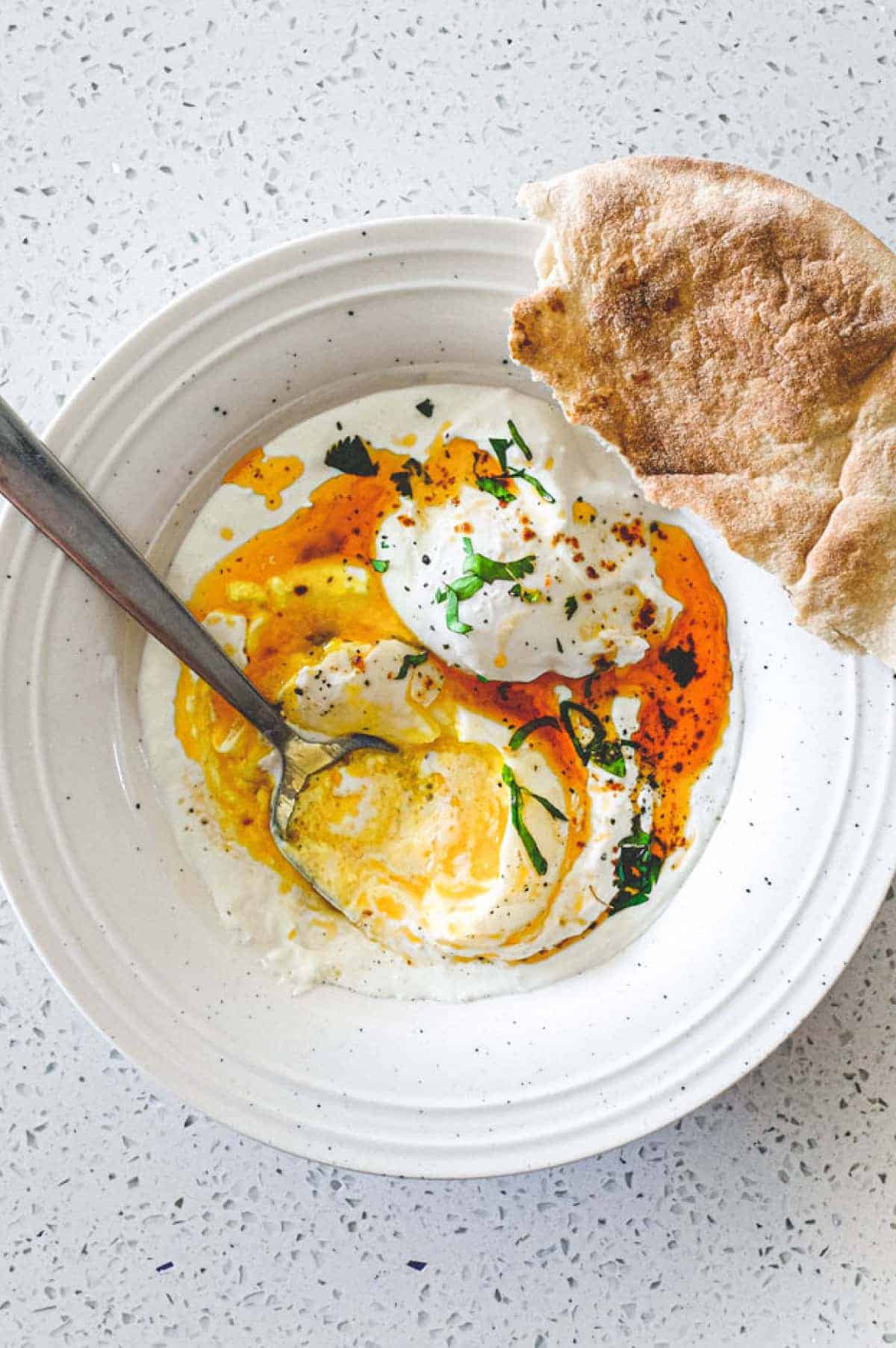 A small bowl of Turkish Cilbir (poached eggs) drizzled in orange Aleppo pepper butter with a piece of flatbread