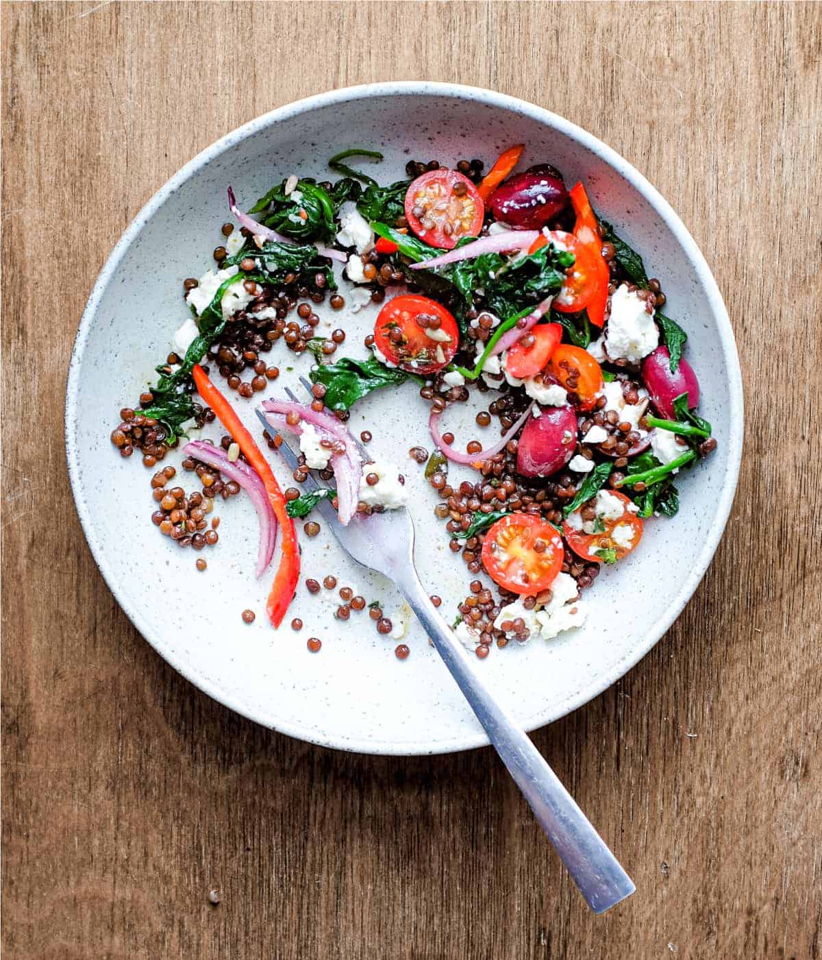 A bowl of Greek lentil salad featuring tomatoes, spinach, lentils, feta and red onion with a fork