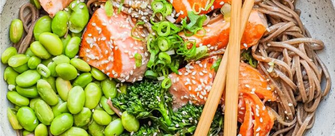 A bowl of salmon with soba noodles with broccolini and edamame with crossed chopsticks.