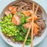 A bowl of salmon with soba noodles with broccolini and edamame with crossed chopsticks.