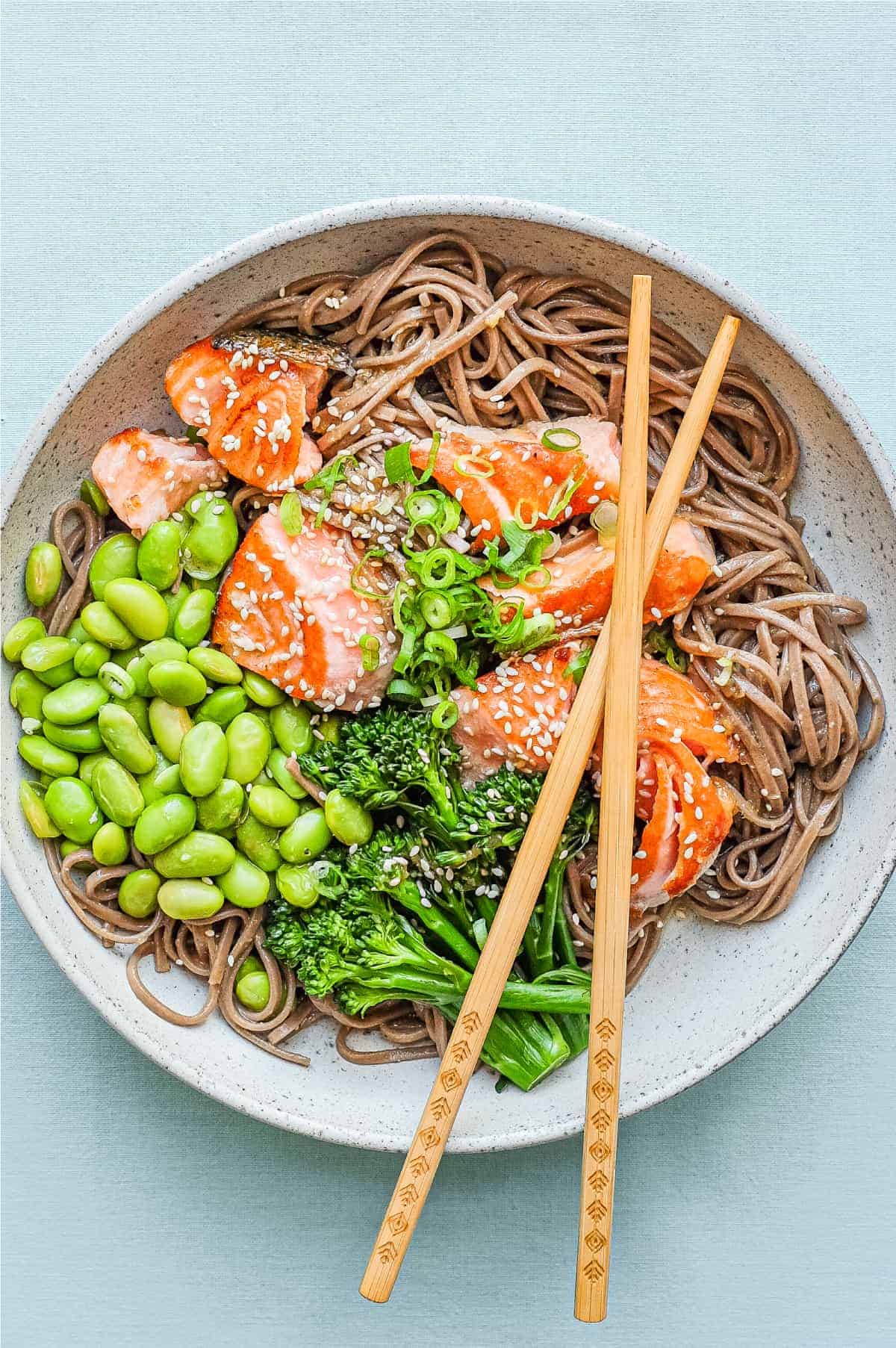 A bowl of salmon with soba noodles with broccolini and edamame with crossed chopsticks.