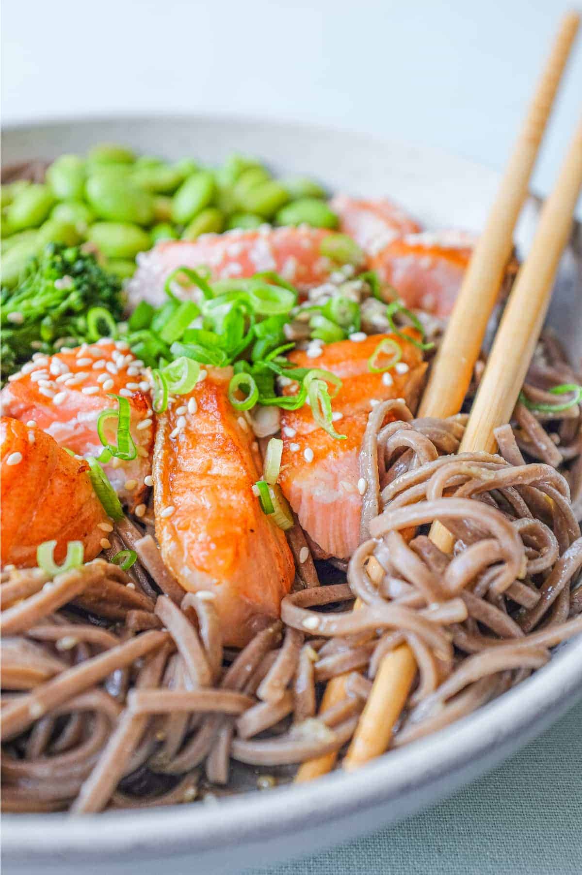 A bowl of salmon with soba noodles with broccolini and edamame with chopsticks