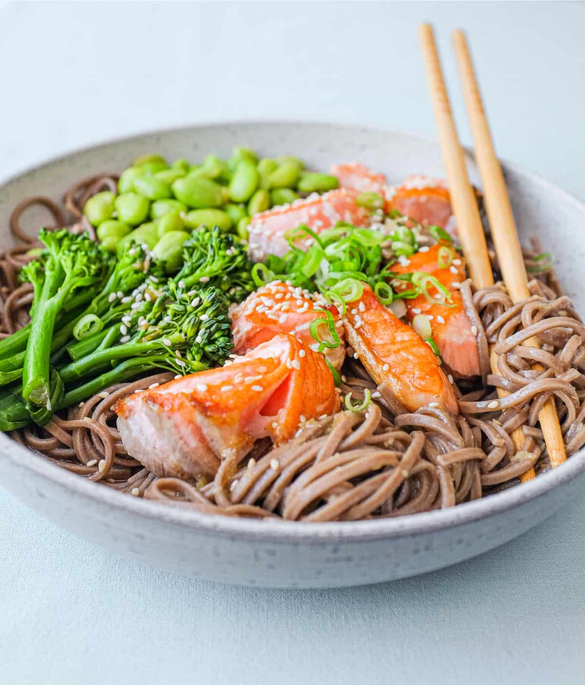 A bowl of salmon with soba noodles with broccolini and edamame with chopsticks