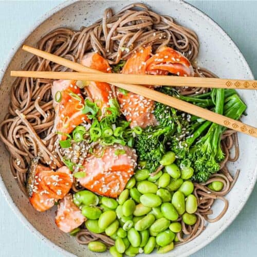 A bowl of salmon with soba noodles with broccolini and edamame with chopsticks