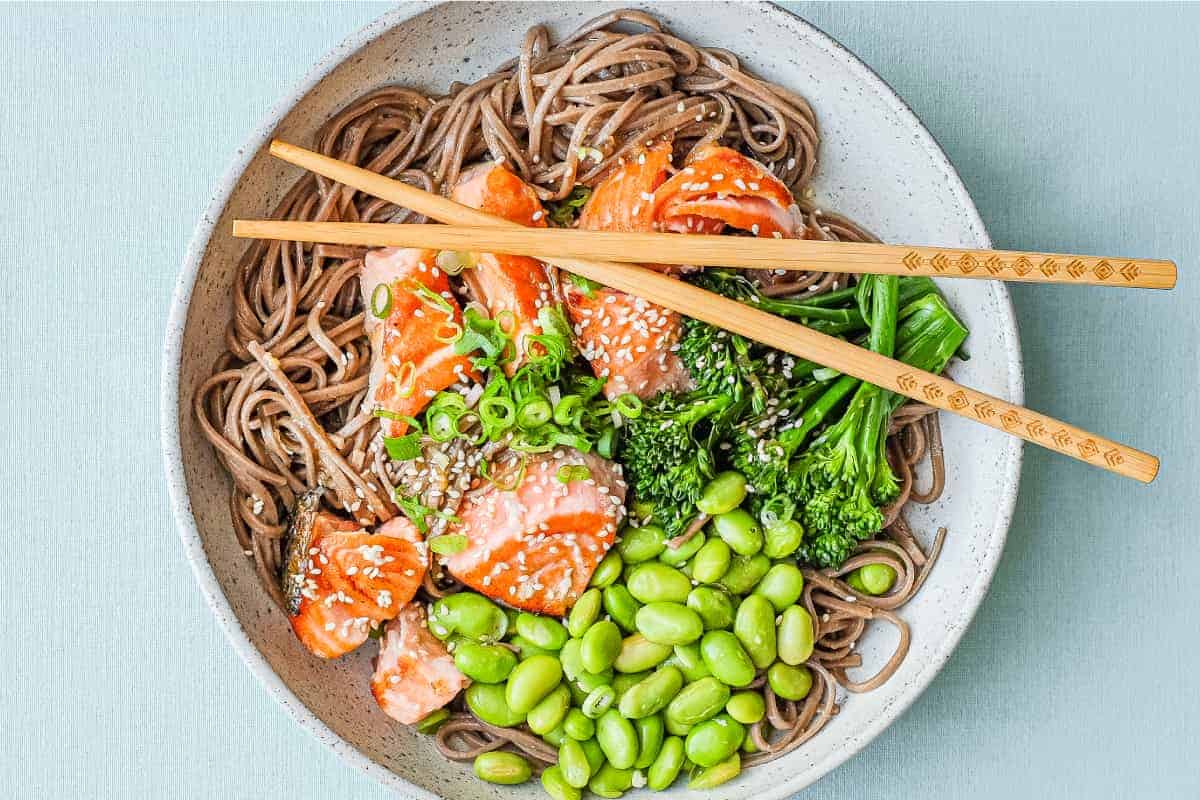 A bowl of salmon with soba noodles with broccolini and edamame with chopsticks