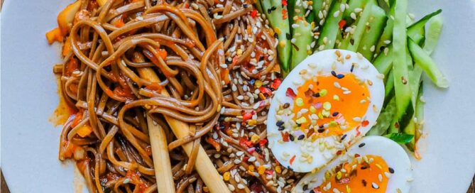 A bowl of Asian noodles called Bibim Guksu. Garnished with eggs and cucumber. Chopsticks are wrapped in the noodles.