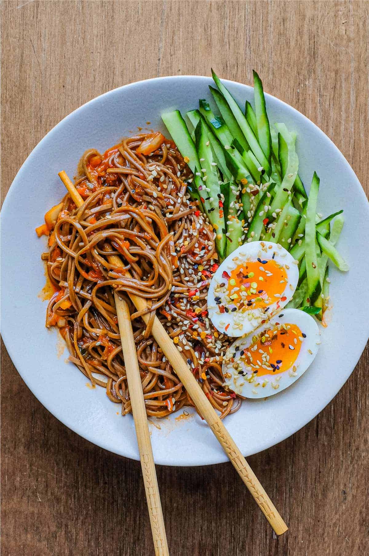 A bowl of Asian noodles called Bibim Guksu. Garnished with eggs and cucumber. Chopsticks are wrapped in the noodles.