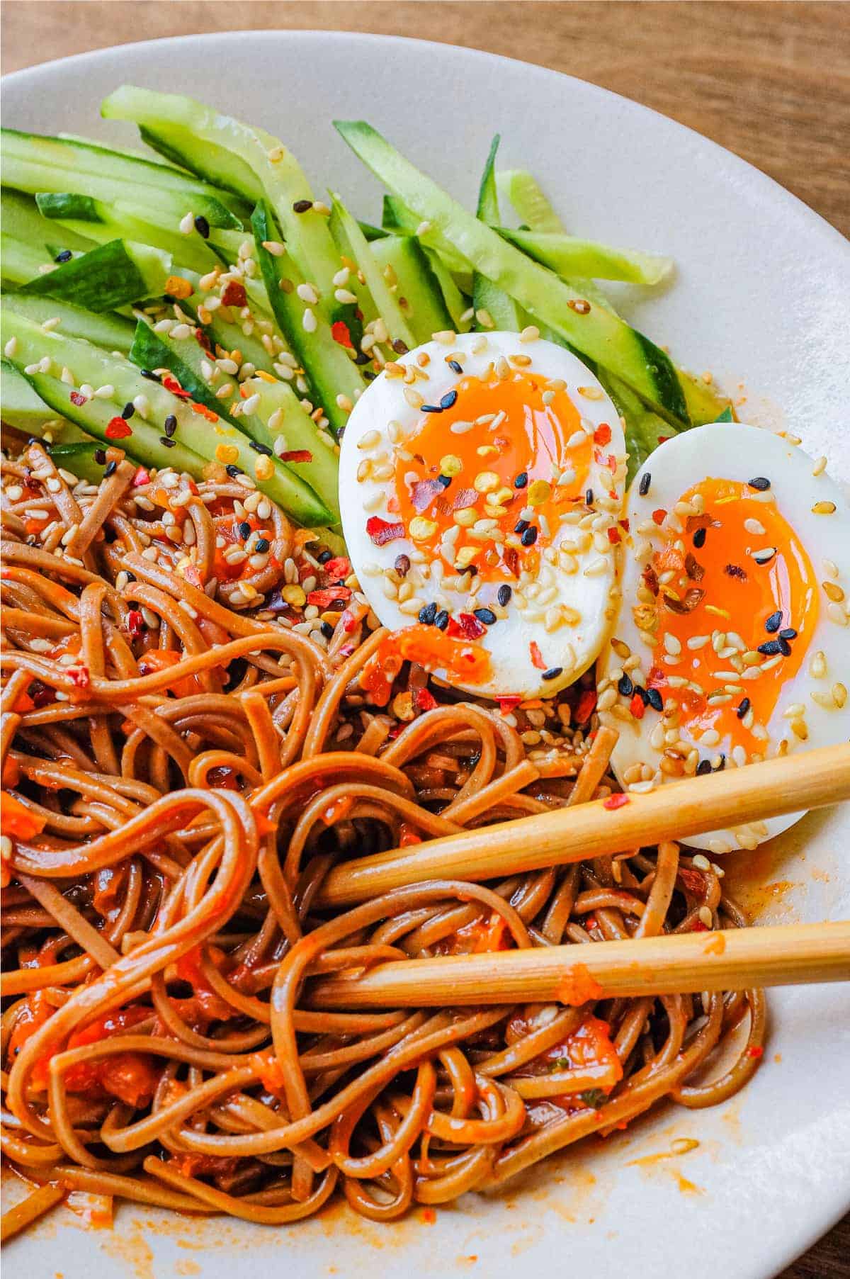 A bowl of Asian noodles called Bibim Guksu. Garnished with eggs and cucumber. Chopsticks are wrapped in the noodles.