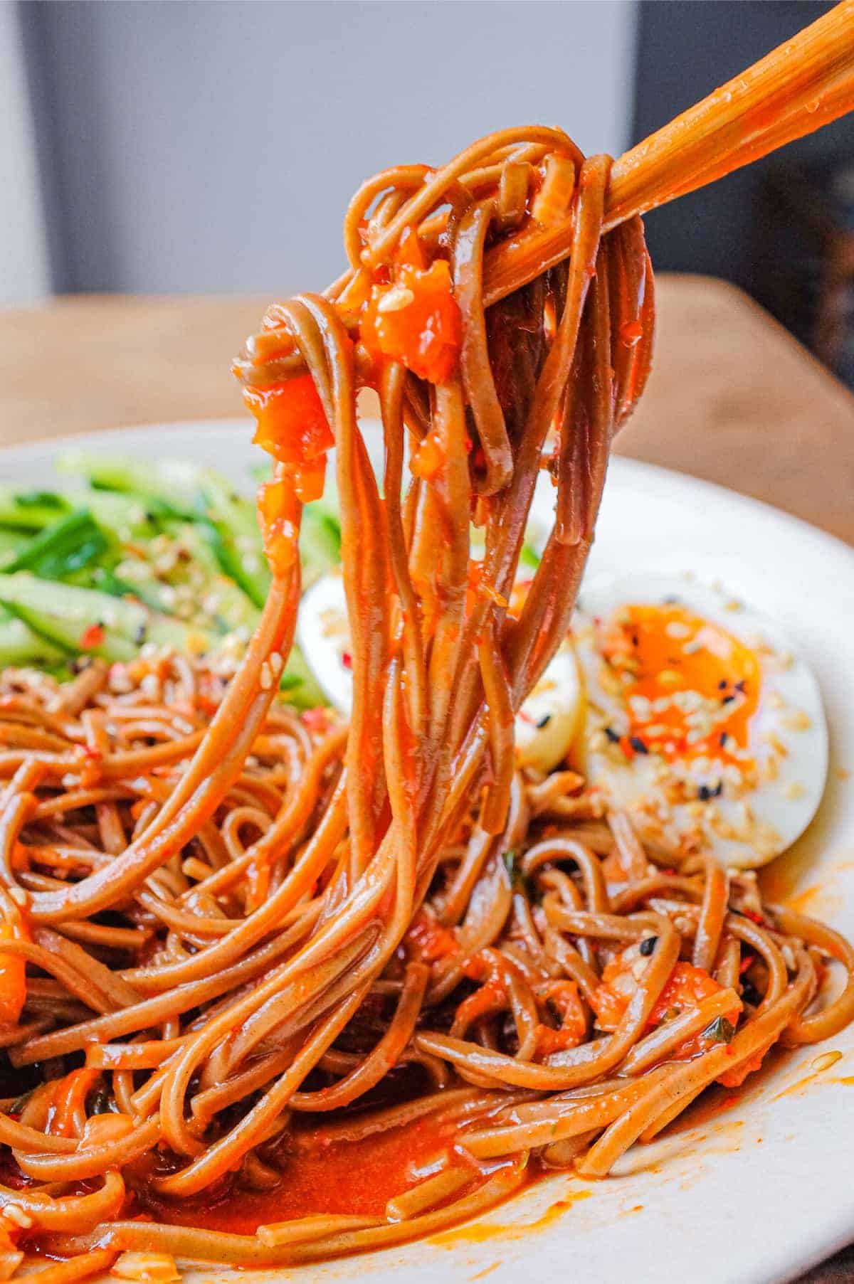 A bowl of Asian noodles called Bibim Guksu. Garnished with eggs and cucumber. Chopsticks hold up the noodles.