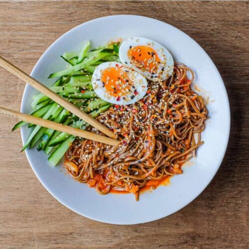 A bowl of Asian noodles called Bibim Guksu. Garnished with eggs and cucumber. Chopsticks are wrapped in the noodles.
