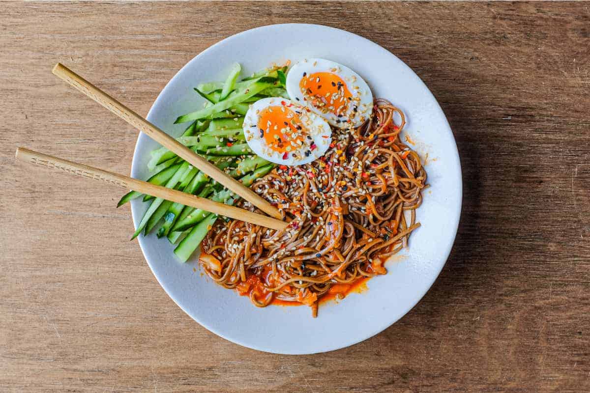 A bowl of Asian noodles called Bibim Guksu. Garnished with eggs and cucumber. Chopsticks are wrapped in the noodles.