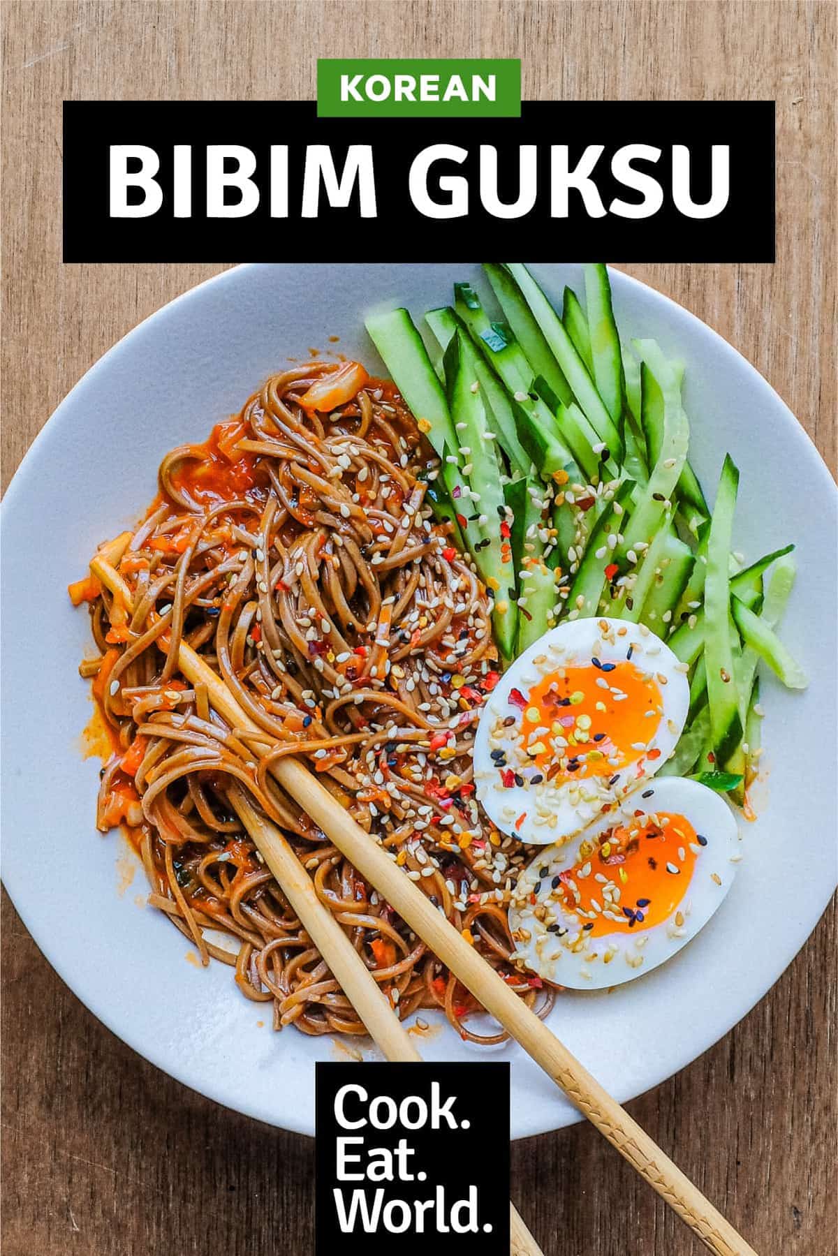 A bowl of Asian noodles called Bibim Guksu. Garnished with eggs and cucumber. Chopsticks are wrapped in the noodles.