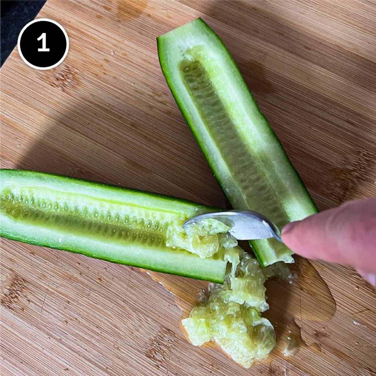 A halved cucumber. The seeds are being removed with a small spoon.