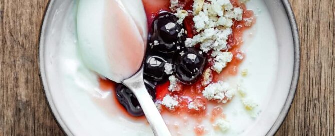 A bowl of lemon cream dessert topped with maraschino cherries and crumbled amaretti biscuits. A spoonful of the cream sits on top.