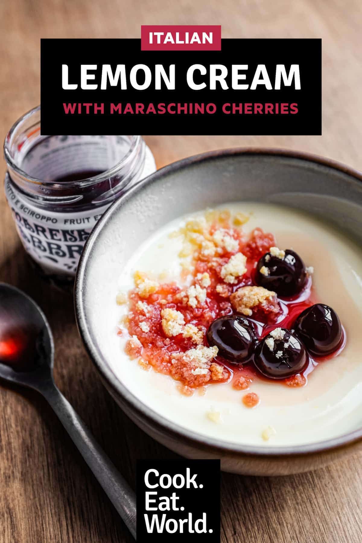 A bowl of lemon cream dessert topped with maraschino cherries and crumbled amaretti biscuits. A small jar of Maraschino cherries and a spoon sit alongside.