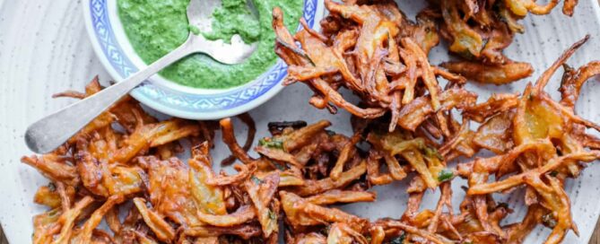 Fried onion pakodas on a platter with a small bowl of coriander chutney and a small spoon.