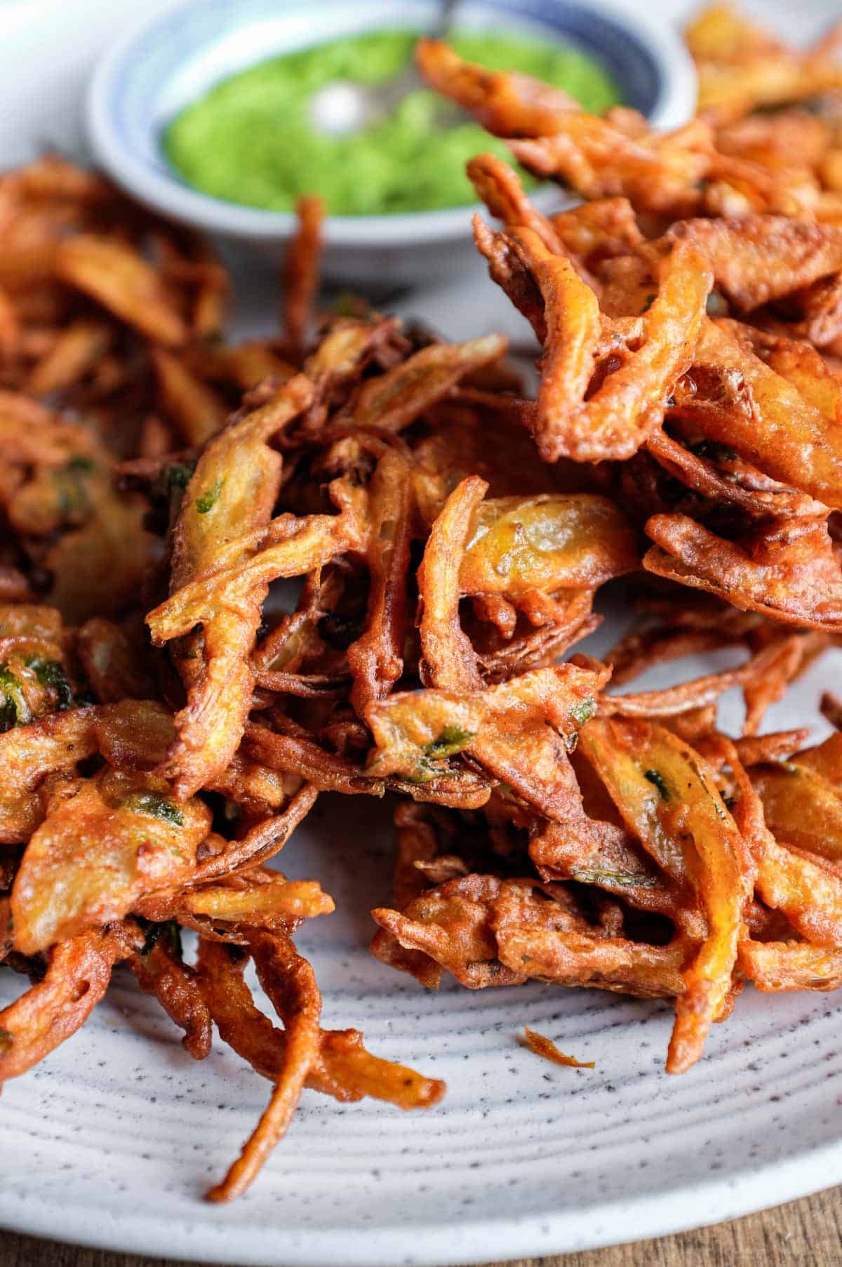 Fried onion pakodas on a platter with a small bowl of coriander chutney and a small spoon.