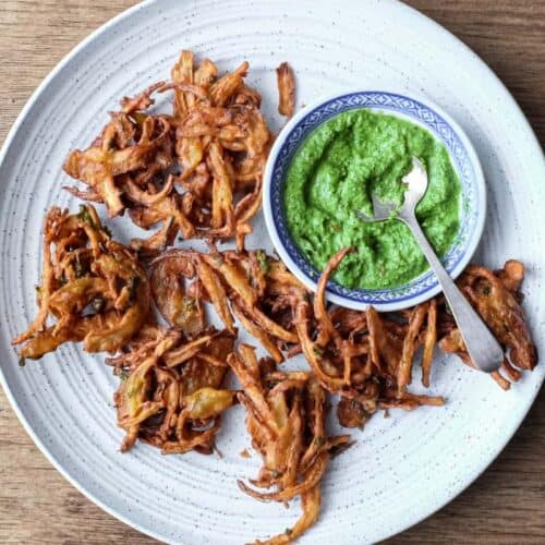 Fried onion pakodas on a platter with a small bowl of coriander chutney and a small spoon.