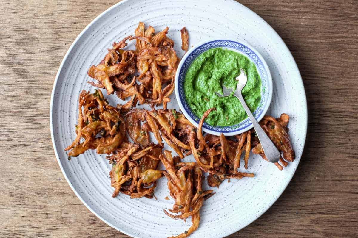 Fried onion pakodas on a platter with a small bowl of coriander chutney and a small spoon.