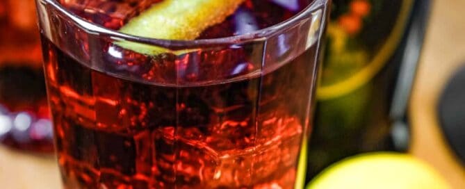 Two cut glass highball glasses of a cocktail called Picon Punch sit alongside a bottle of Picon Amer and a lemon.
