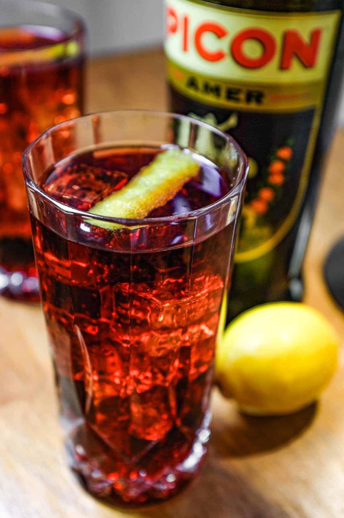 Two cut glass highball glasses of a cocktail called Picon Punch sit alongside a bottle of Picon Amer and a lemon.