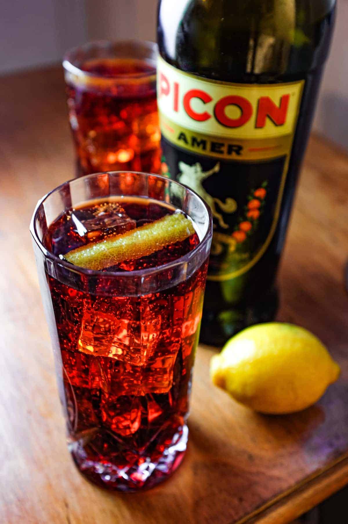 Two cut glass highball glasses of a cocktail called Picon Punch sit alongside a bottle of Picon Amer and a lemon.