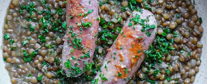 A shallow bowl has a layer of creamy lentils, topped with two Italian pork and fennel sausages. The dish is garnished with fresh parsley.
